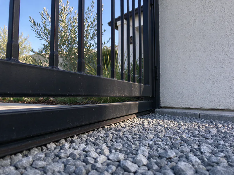 Close view of a steel gate bottom just above a gravel strip beside a stucco wall.